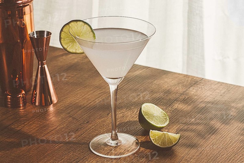 A clear cocktail sits in a glass with a lime slice on the rim. A copper shaker and halved lime are also on the wooden table. Bright light comes in from the window