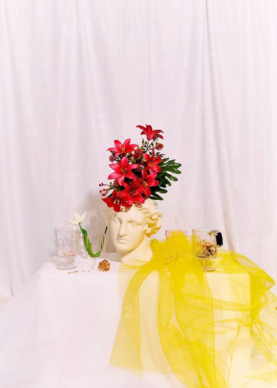 A classical white sculpture head is decorated with vivid red flowers, set against a soft white backdrop. Nearby, glassware and a yellow fabric add a touch of elegance