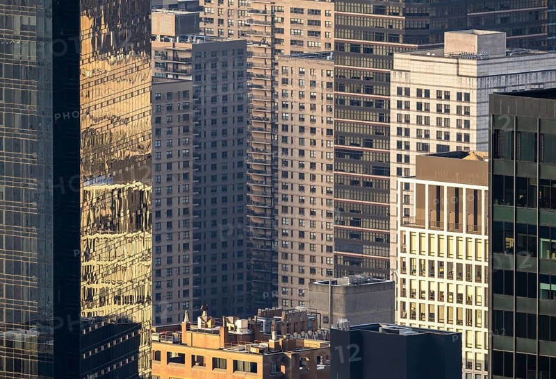 From above, the sunlit facade of New York's high rise buildings and skyscrapers showcases an urban texture of windows and reflections. The intricate play of light highlights the dense architectural forest in the heart of the city