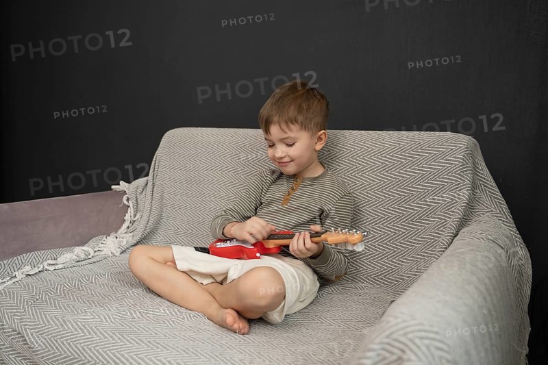 A young boy sits on a cozy sofa, playing a small red toy guitar. His relaxed posture and focused expression reflect joy and creativity in a comfortable home setting