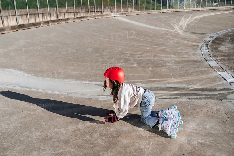 A young girl with a red helmet and protective gloves cautiously gets up from the ground, from a fall during roller skating practices, showing determination, at an outdoor skate park