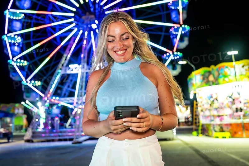 Young blonde woman smiling while texting on a smartphone at an amusement park or fair, surrounded by colorful neon lights from a ferris wheel and other attractions at night