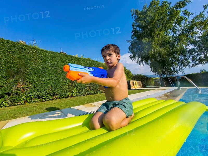 Young boy on a green inflatable float in a sunny pool, squirting a water toy and laughing as he splashes and enjoys a carefree summer day outdoors
