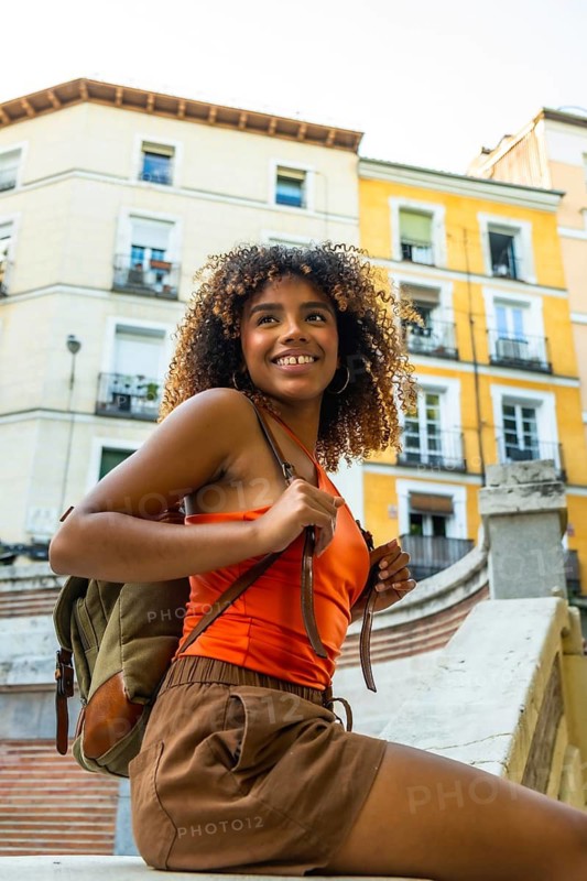 Young woman with curly hair and a backpack smiling while exploring an urban street during summer, experiencing freedom, adventure and joy in her journey