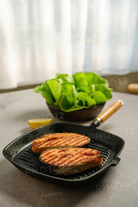 Grilled salmon steaks on frying pan on kitchen tabletop. Bowl of fresh salad sits on the table. Soft light comes from a window. A lemon slice is placed next to frying pan