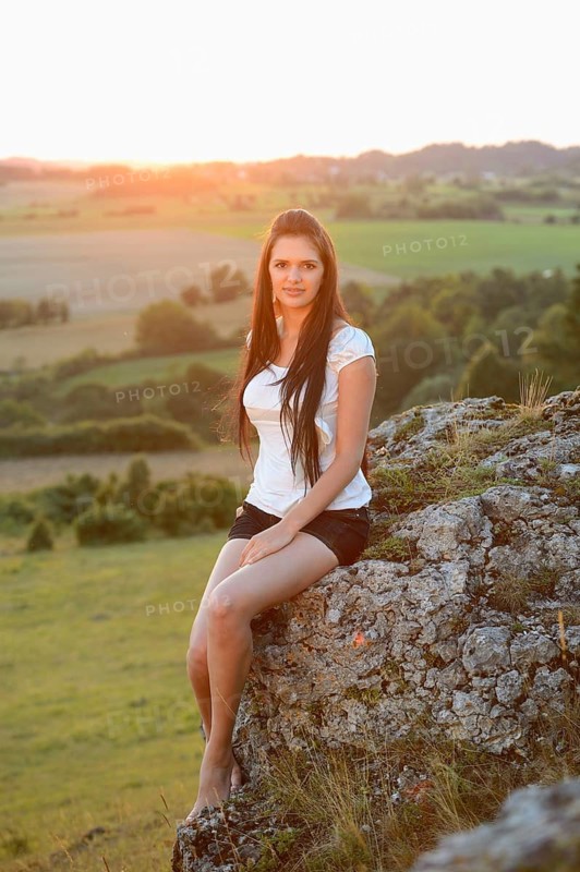 A woman with long hair sits on a rock in nature during sunset, wearing a white shirt, Bavaria