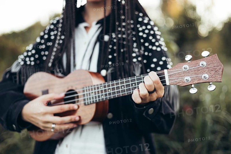 A woman in a black jacket plays a wooden ukulele against a blurred backdrop of grass during sunset, creating a warm, calming atmosphere that encapsulates the joy of music