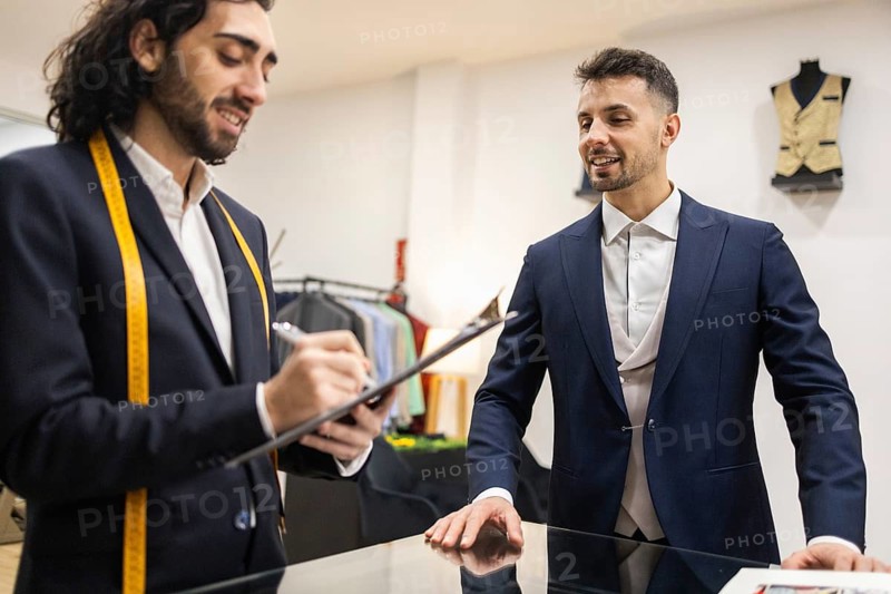 A tailor consults with a client in a suit store, taking notes while discussing suit options. They stand at a counter surrounded by outfits and measuring tools
