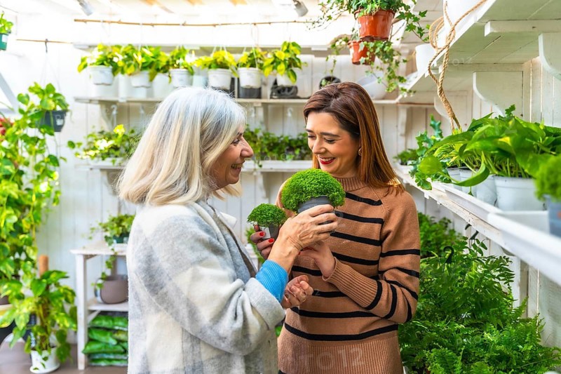 Senior woman and her daughter smiling and choosing green potted plants together in a bright garden center, sharing a hobby and enjoying time together among lush indoor greenery