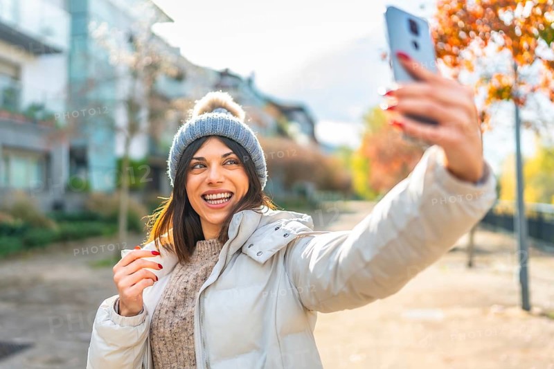 Happy beauty woman smiling at camera while taking selfie with smartphone standing in the street in autumn