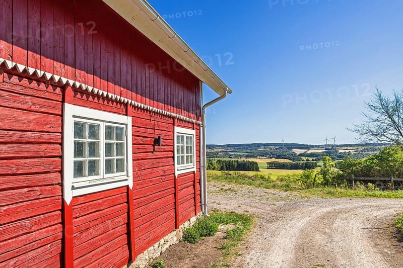 Red wooden barn with white trim stands beside a curved dirt road in a scenic rural landscape with green fields under a clear blue sky, Sweden