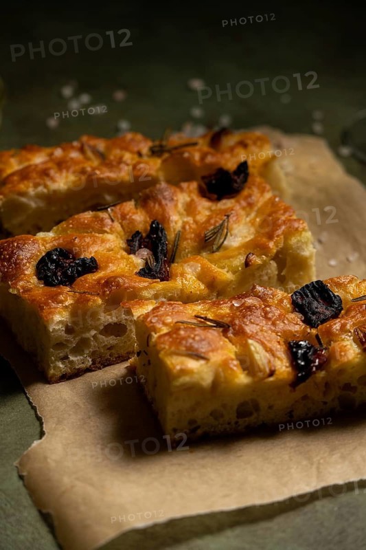 Close up of freshly baked homemade focaccia bread, topped with rosemary and black olives, placed on parchment paper. The golden crust and soft texture showcase its delicious appeal