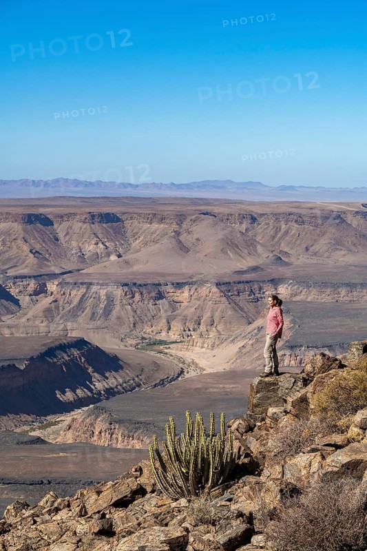 Woman standing on a rock at the edge of the precipice in front of the gorge landscape of the Fish River Canyon, Fish River Gorge, Namibia