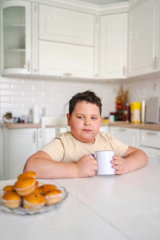 A child holds a mug at a kitchen table with muffins nearby. The bright kitchen background creates a cozy atmosphere, symbolizing a pleasant breakfast moment