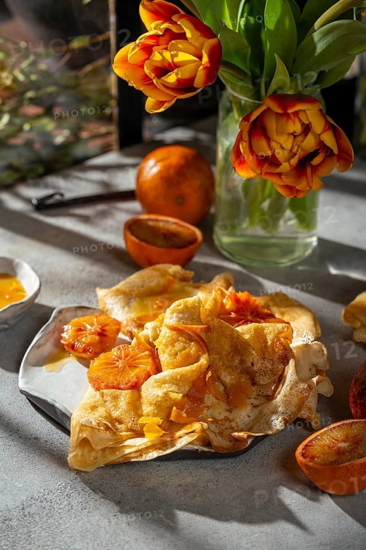 A plate of crepes Suzette garnished with caramelized slices of blood orange in a rich butter sugar sauce. The setting includes fresh tulips and a rustic tabletop creating a cozy, inviting atmosphere