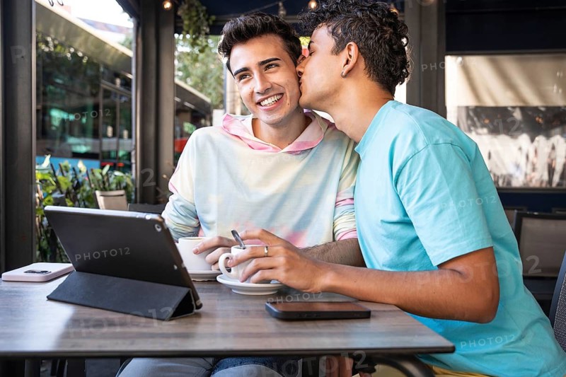 A joyful gay couple shares coffee and a tender moment at an outdoor cafe. One leans in for a kiss, both smiling, expressing love and connection amid a lively urban setting