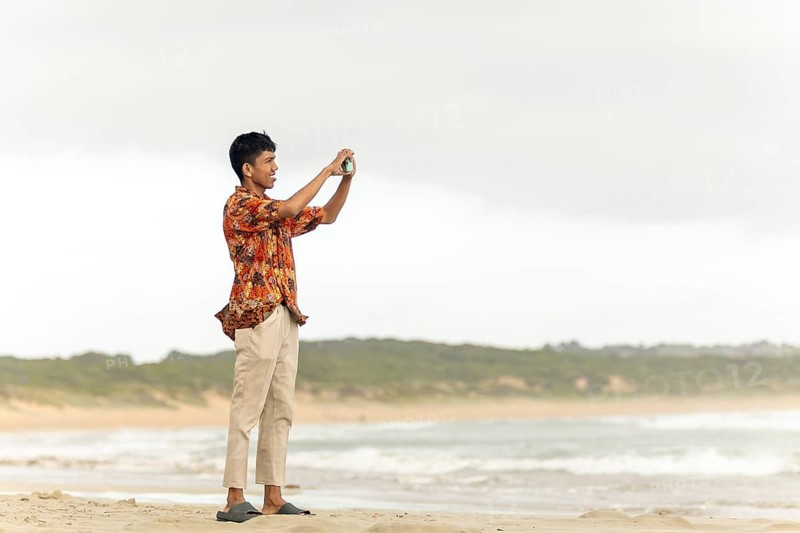 An indonesian man wearing traditional batik clothing captures a scenic moment on an Australian beach, blending cultural heritage with a vibrant coastal landscape
