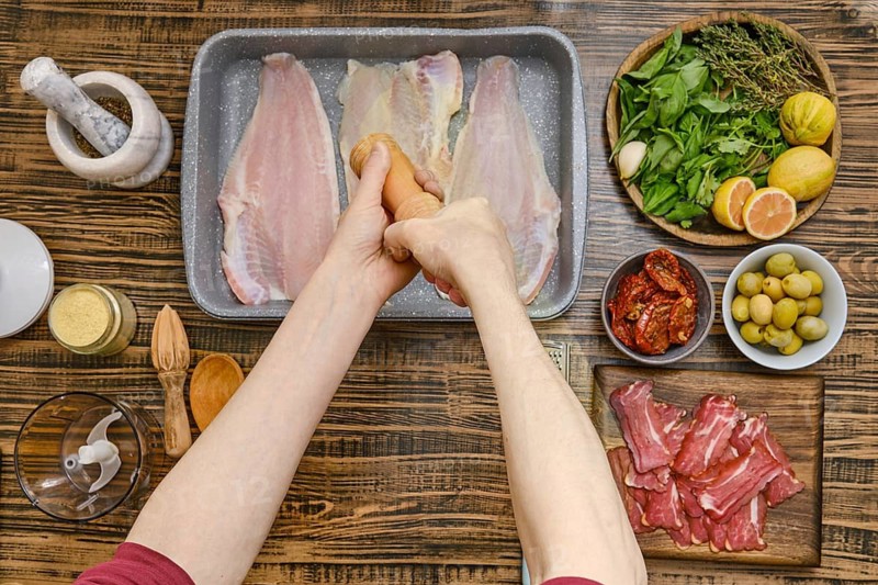Hands sprinkle fish fillet with salt while surrounded by fresh herbs, olives, and sliced meats
