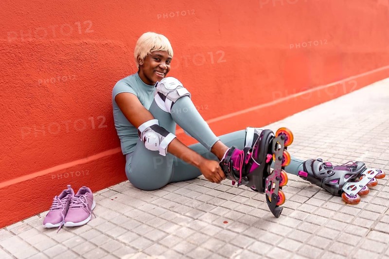 Happy african sportive woman putting on inline skates sitting in the street leaning on red facade