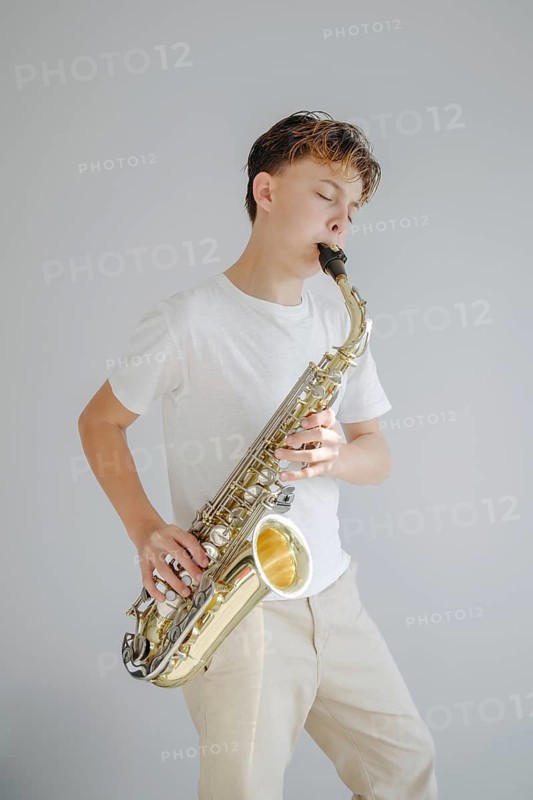 A young boy musician passionately plays the saxophone, eyes closed, dressed in casual attire. The neutral background highlights his focus and dedication to music