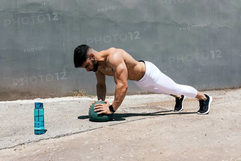 Athletic man outdoors performing a plank exercise using a medicine ball. He is showcasing strength and balance on a sunny day, emphasizing fitness and health