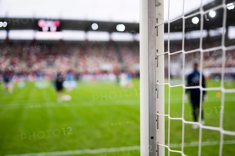 Close-up of a white soccer goalpost and net on a green grass pitch with blurred players training in a stadium background in Lubin, Poland