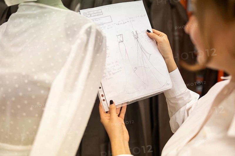 A fashion designer holds and reviews detailed sketches in a atelier, focusing on the development of a garment. A polka dot mannequin is visible in the background, enhancing the creative atmosphere