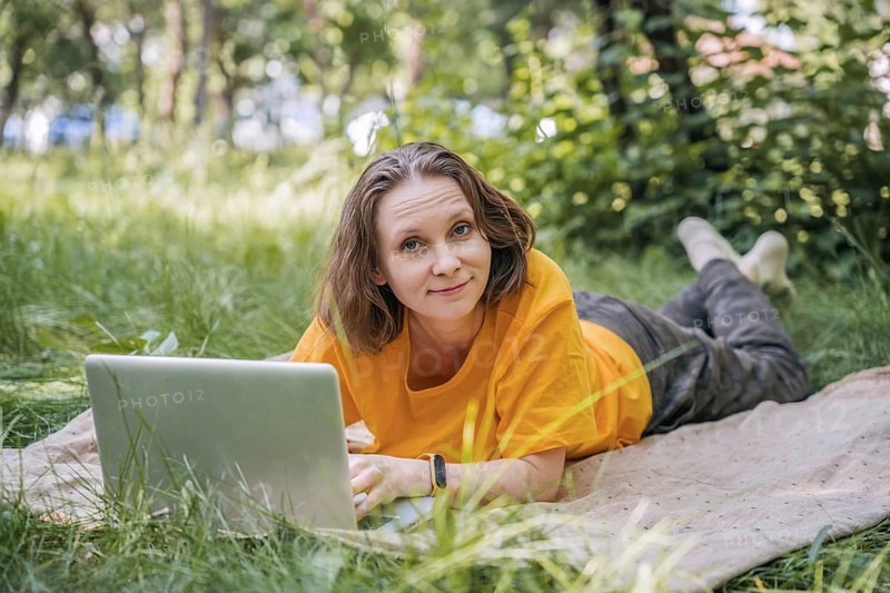 A woman is lying on a blanket in the park, working on her laptop. The serene natural environment provides a refreshing setting for remote work, blending technology with nature