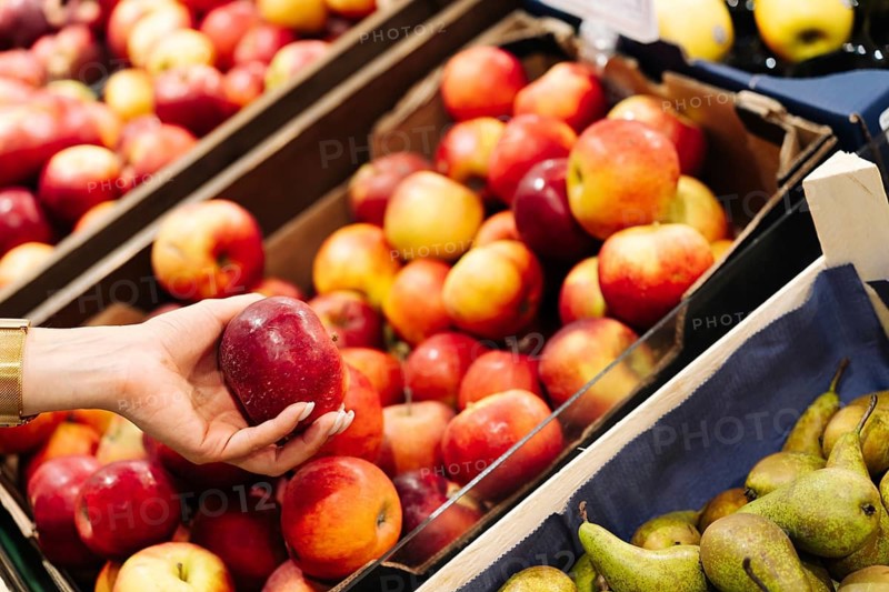 A hand holds a red apple above a display filled with assorted apples and pears. The scene highlights fruit variety, produce comparison, and the process of choosing fresh items at a market