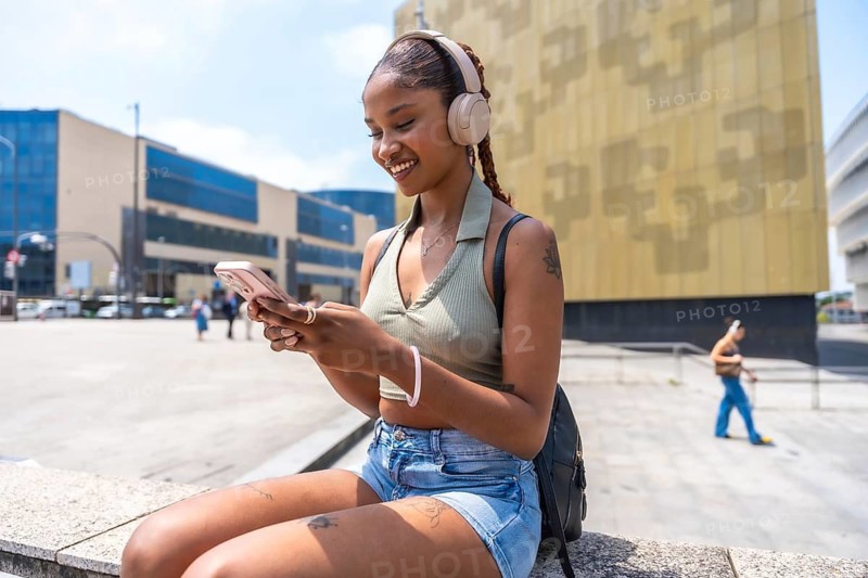 Young woman smiling while using a smartphone and listening to music through headphones, enjoying leisure time in an urban city environment on a sunny day