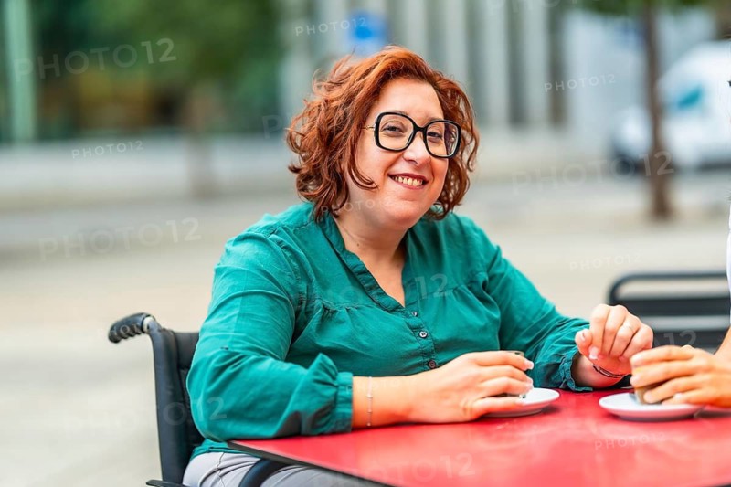 Woman with disability using electric wheelchair drinking coffee with friend sitting in an outdoor cafeteria in the city