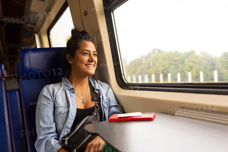A cheerful woman enjoys a scenic train ride, gazing out the window at lush green landscapes A red smartphone rests on the table The scene captures relaxation and adventure
