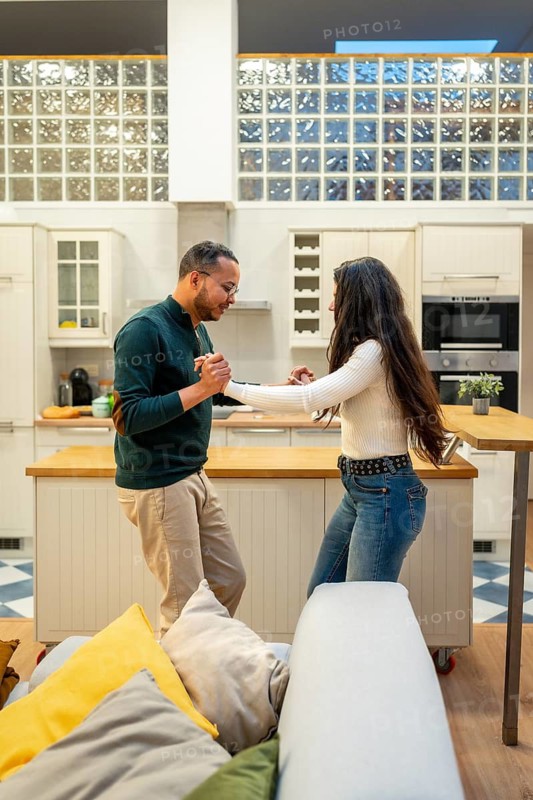 Young multi ethnic couple smiling and holding hands while dancing in a modern open plan kitchen and living room, sharing a joyful, affectionate moment at home