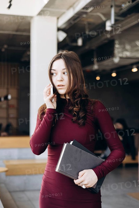 A young female student stands indoors, balancing books while talking on her phone. Her expression suggests engagement and focus as she converses in a modern setting