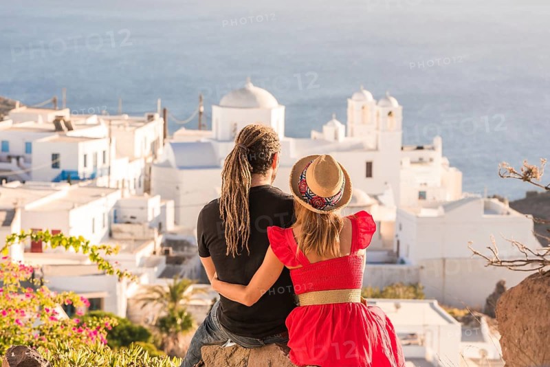 A couple embraces while admiring the stunning ocean view and traditional white architecture of Greek. Their casual attire and joyful vibe capture the essence of travel romance in Plaka, Milos island
