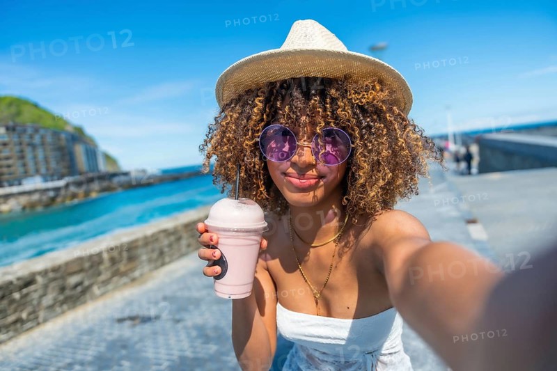 Cool woman with curly hair and hat taking a selfie drinking milkshake next to the sea
