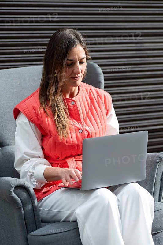 A fashion woman in casual attire works on her laptop while seated in a comfy armchair. She appears focused, reflecting a cozy work environment