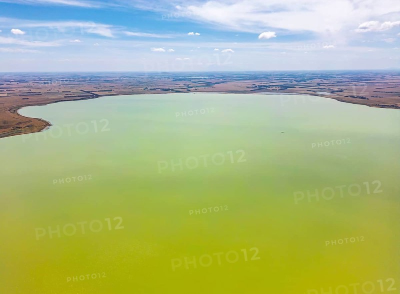 Aerial photograph capturing a vast, calm green lake surrounded by expansive, flat countryside in Victoria, Australia. The blue sky with scattered clouds adds serenity to the tranquil scene