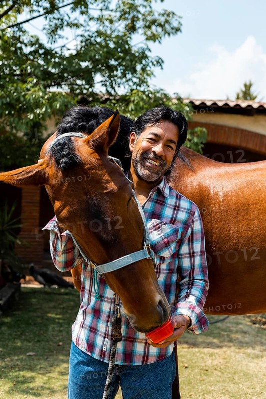 A man in a plaid shirt shares a joyful moment with his horse in a sunny garden. The horse nuzzles affectionately while the man smiles, showcasing a warm human-animal bond