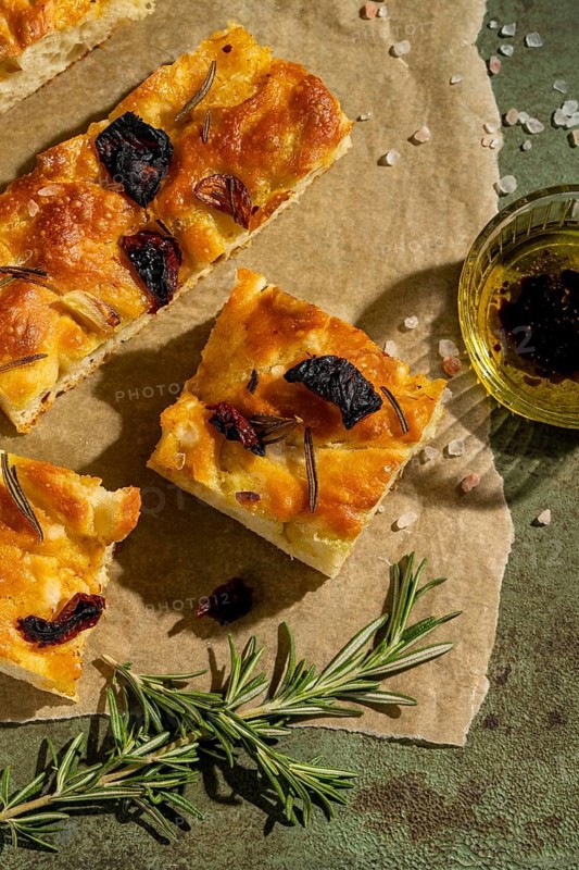 Freshly baked homemade focaccia slices garnished with rosemary and sun dried tomatoes on parchment paper, accompanied by a sprig of rosemary and a small bowl of olive oil