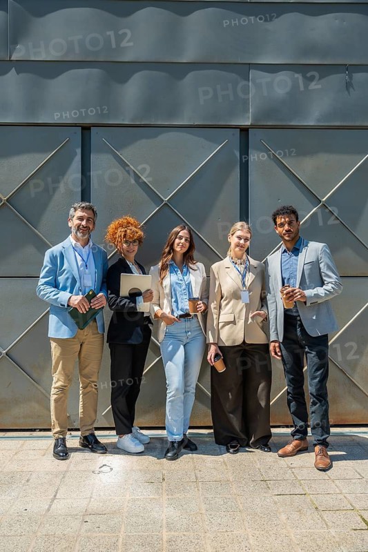 Diverse business professionals standing outdoors in front of a modern building, smiling at the camera and holding coffee cups and electronic devices, representing unity, collaboration, and success