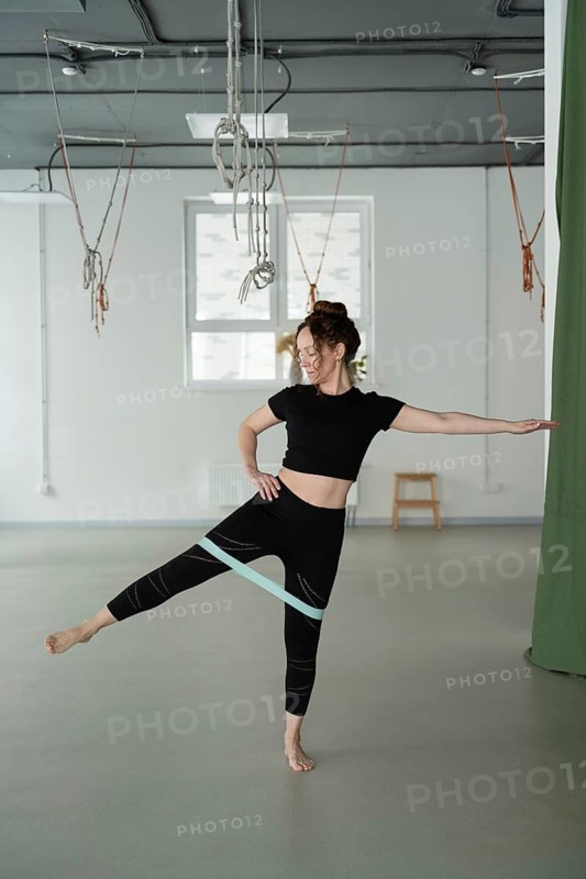 A woman in athletic wear performs a yoga exercise using a resistance band in a studio. The setting is calm, with suspended equipment in the background