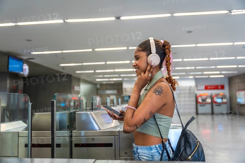 Young black woman listening to music with headphones and smartphone, smiling while passing through turnstile at a modern subway station, experiencing daily urban mobility and connectivity