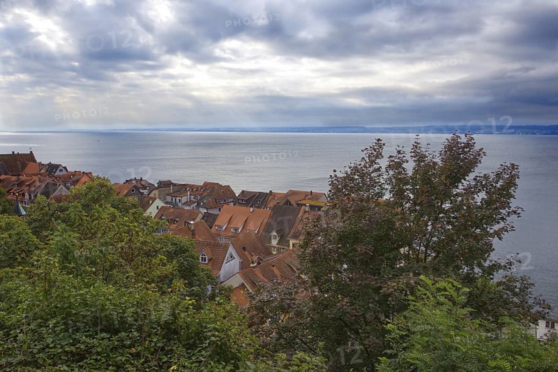 View of the houses in the old town of Meersburg with red tiled roofs. Behind it, Lake Constance with wooded shore in the distance under cloudy skies, Meersburg, Baden-Württemberg