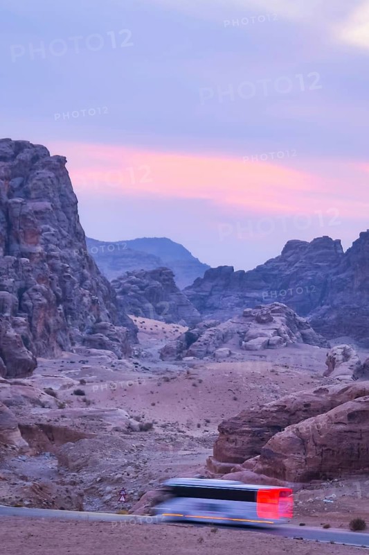 White passenger coach bus driving asphalt road winding through sandstone rock mountains canyon desert landscape at night leading ancient archaeological tourist attraction - Petra Jordan