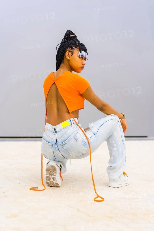 A young trap dancer with braids. Black ethnic girl with orange t-shirt and cowboy pants. Backs of Back Agachada looking seductively