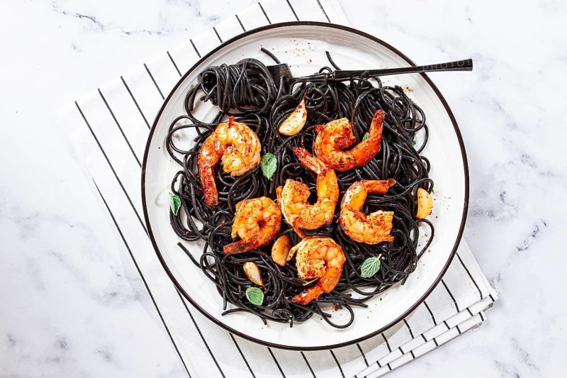 Black spaghetti with shrimp, on a white plate with a fork, on a striped napkin, marble background, homemade, no people