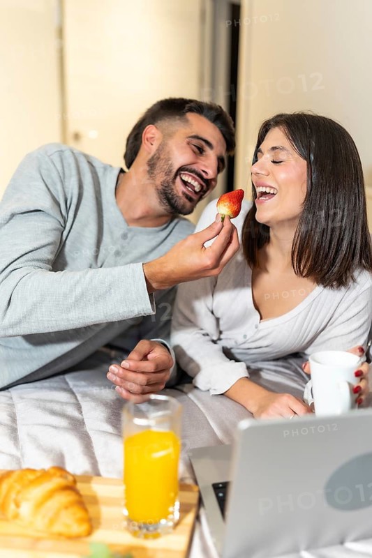 Young man offering a strawberry to his smiling girlfriend during a romantic breakfast in bed, with orange juice, croissant, and a laptop nearby