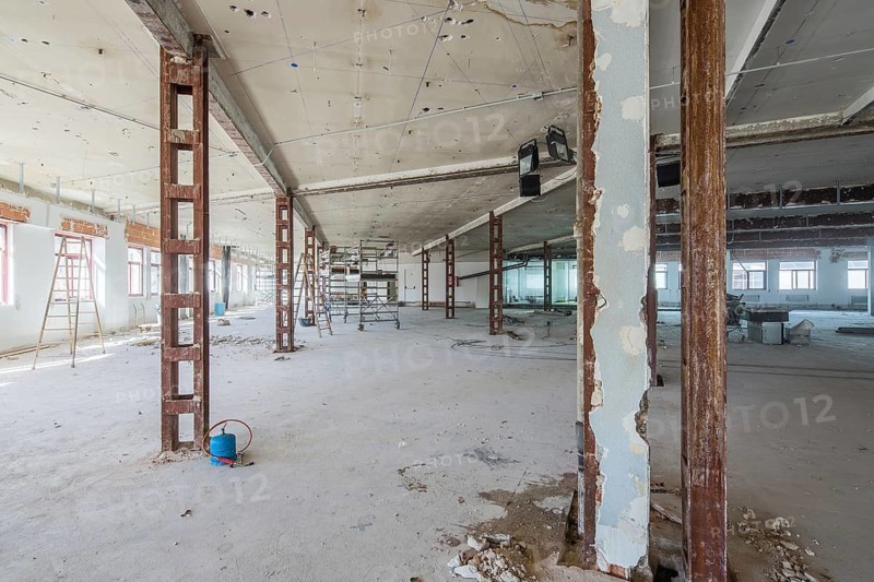 A large space under renovation, showcasing exposed steel beams and construction equipment. Natural light streams through tall windows, illuminating the open area