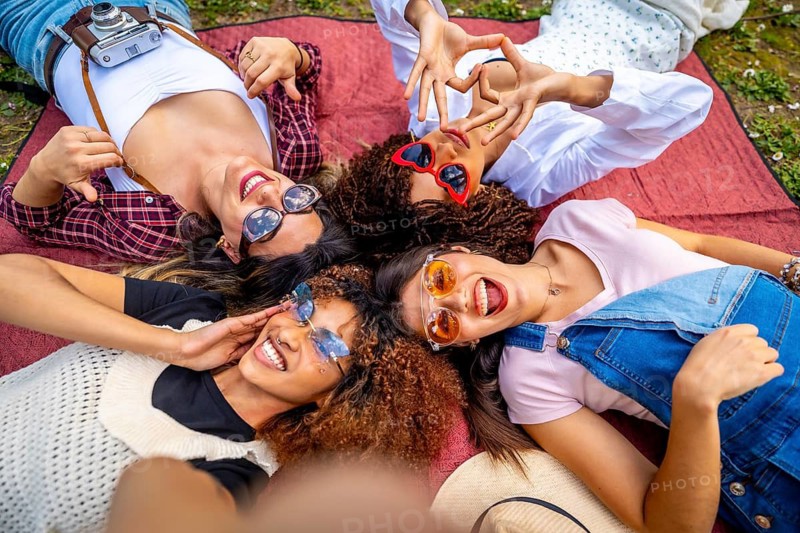 Four young women lying on a blanket and making a heart shape with their hands, taking a selfie and smiling